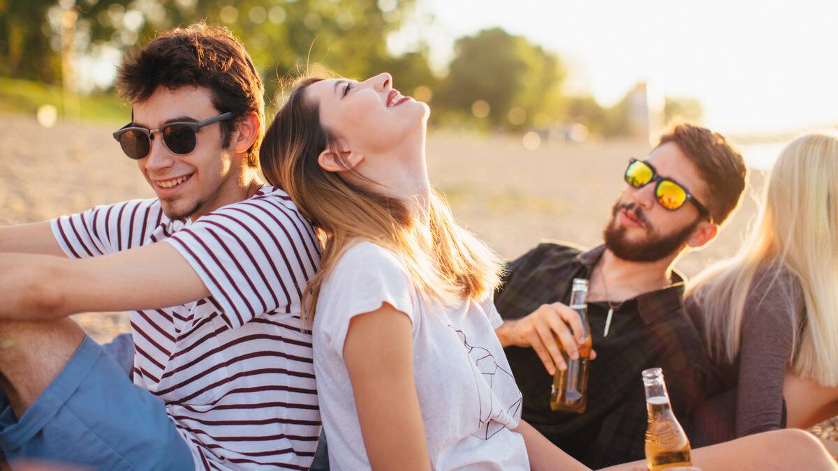 Friends sitting outdoors together, smiling, holding drinks, two with their backs against each other's.