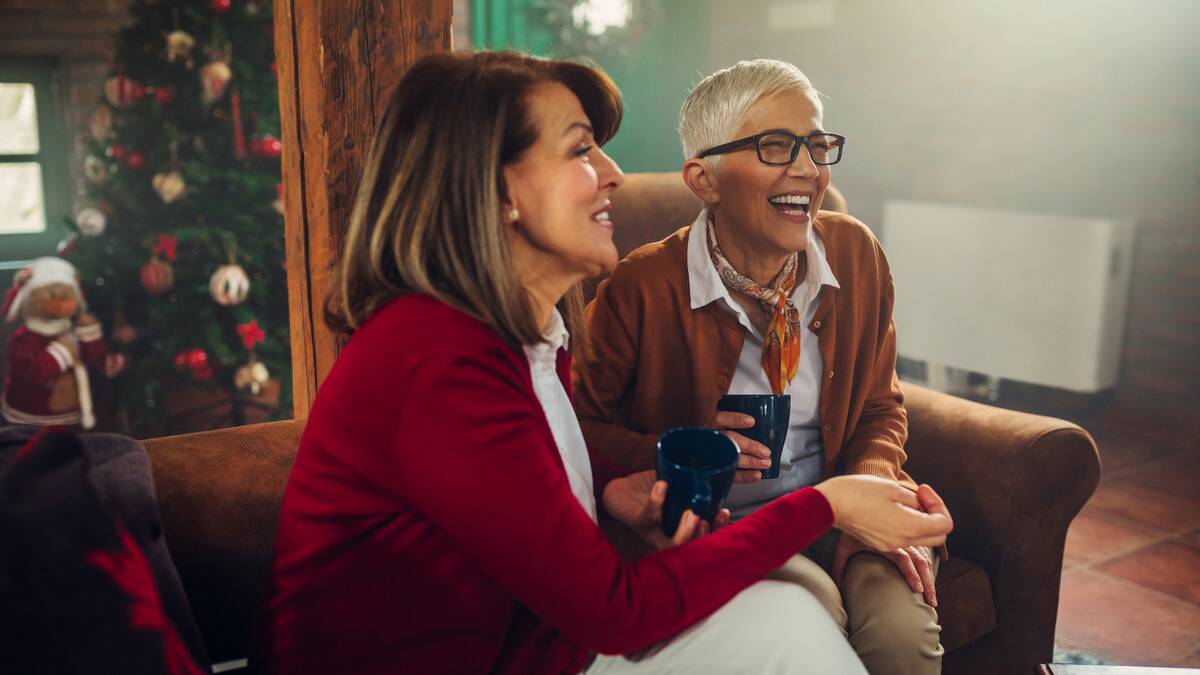 Two women sitting on a couch indoors together, smiling and laughing as they chat.