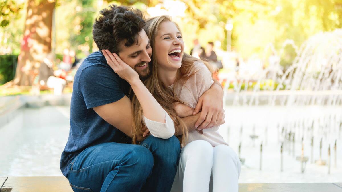 A couple sitting side by side in front of a fountain, the man hugging the woman, the woman cupping his cheek, both smiling and laughing.