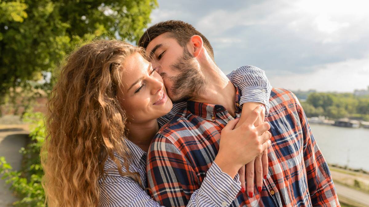 A couple standing outside, the woman hugging the man from behind, the man leaning back to kiss her cheek.