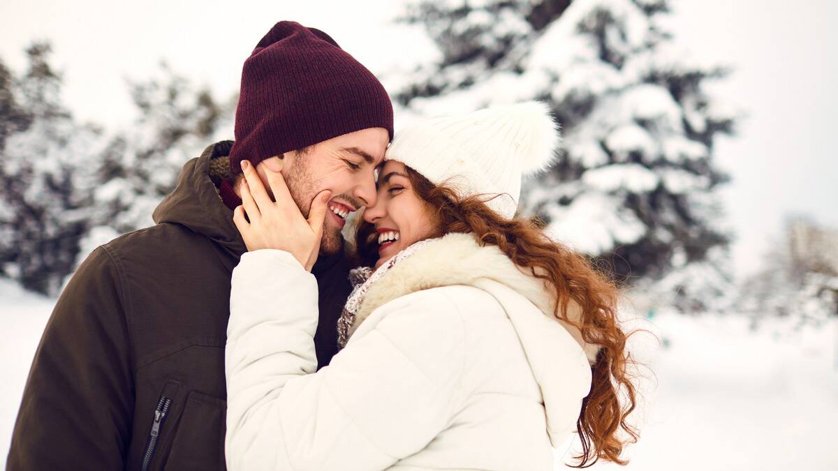 A couple standing outside in the snow, chest to chest, the woman with a hand on the man's cheek, both smiling with their eyes closed.