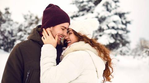 A couple standing outside in the snow, chest to chest, the woman with a hand on the man's cheek, both smiling with their eyes closed.