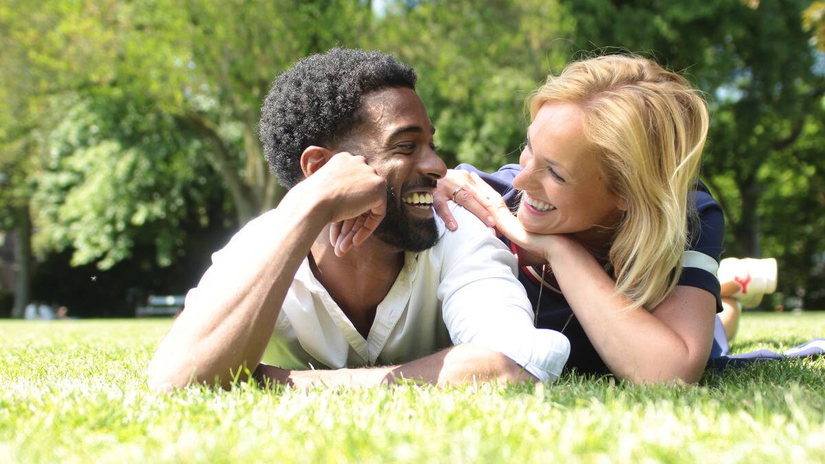 A couple both laying on their stomachs side by side in the grass, propped up on their elbows, smiling at one another.