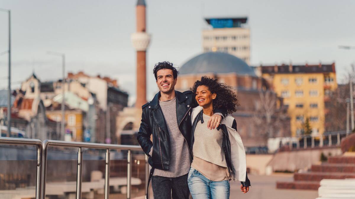 A couple walking down a city street with an arm around each other, smiling.