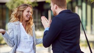 A man pleading to a woman, his hands together, while she shrugs and laughs it off.