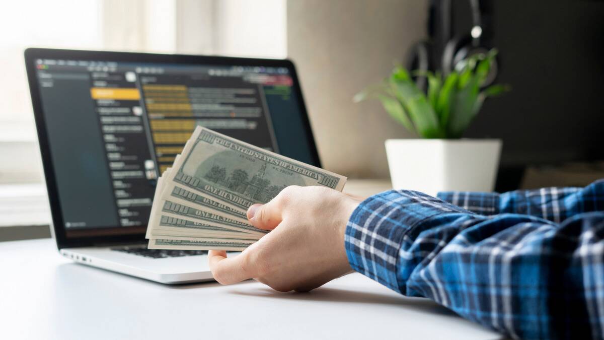 A hand holding a stack of $100 bills in front of a laptop.