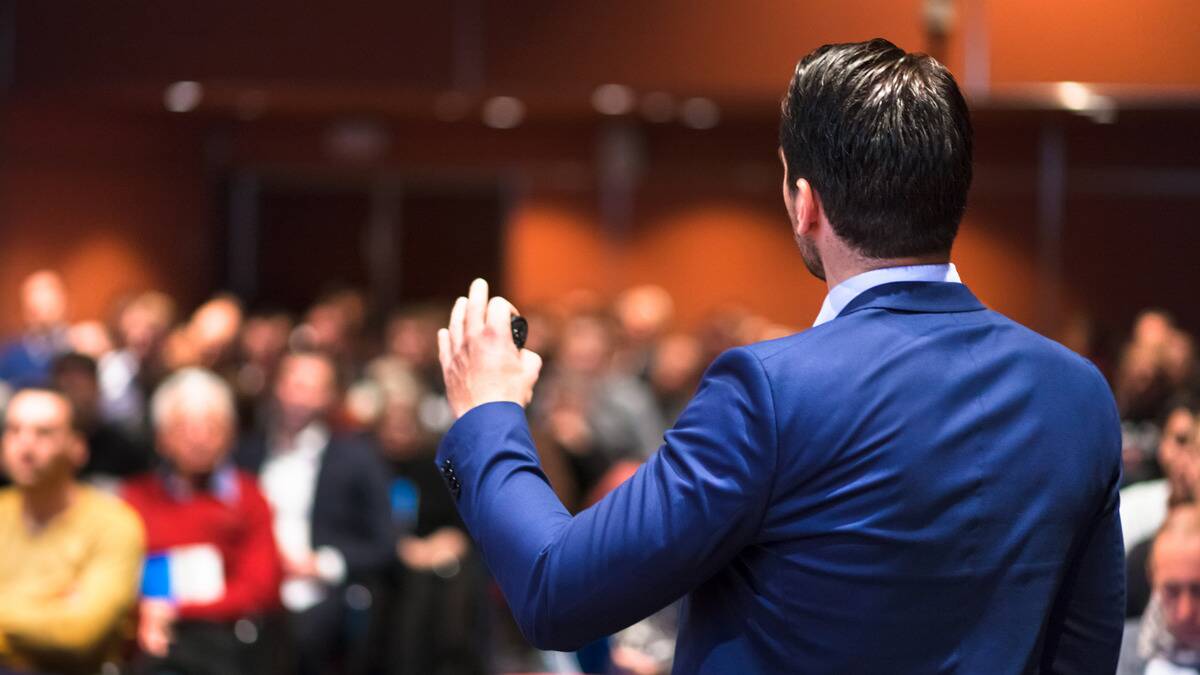 A shot from behind of a man in a suit speaking to a crowd of people.