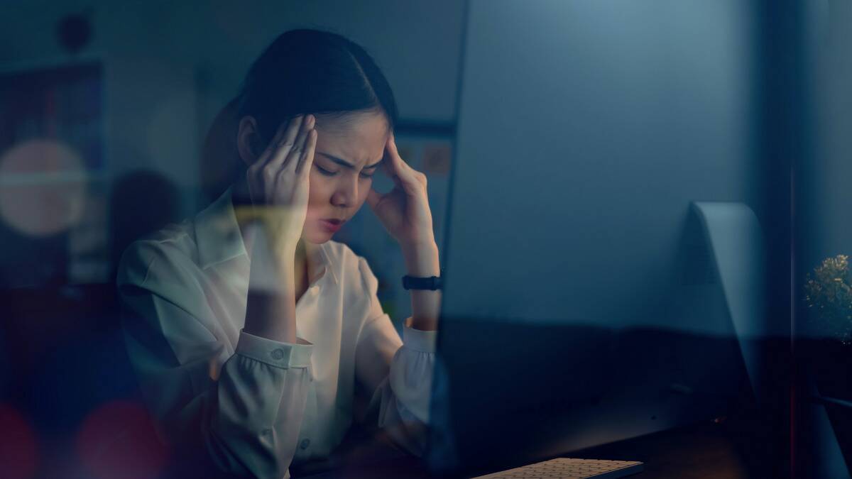 A woman sitting in the dark in front of a computer, both hands against her temples, looking stressed.