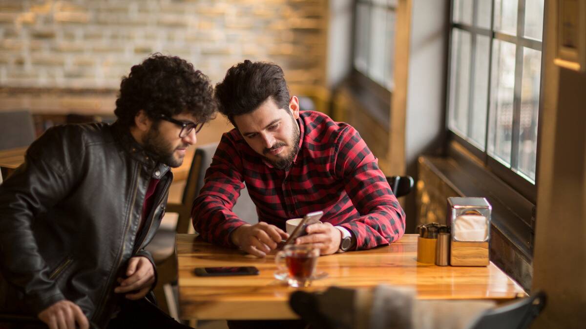 Two friends sitting at a cafe table, one showing the other something on his phone.