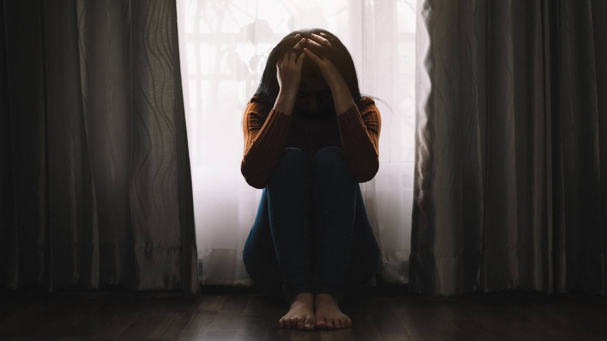 A woman sitting on the floor in front of a window that's being covered by a curtain, knees drawn up, head in her hands.