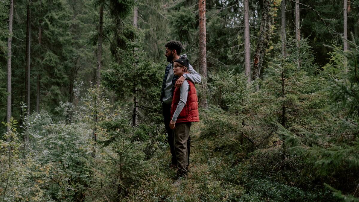 A couple standing in a forest, one with their arm around the other.