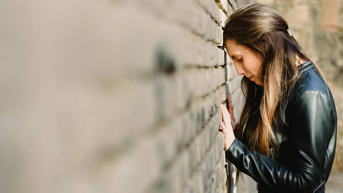 A woman leaning her forehead on a brick wall, her hands against the wall as well, looking down sadly.