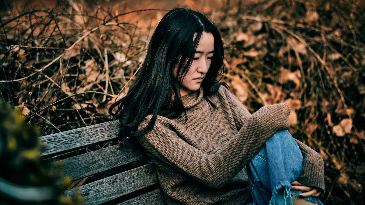 A woman sitting on a bench outside, one knee drawn up, looking sad.