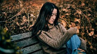 A woman sitting on a bench outside, one knee drawn up, looking sad.