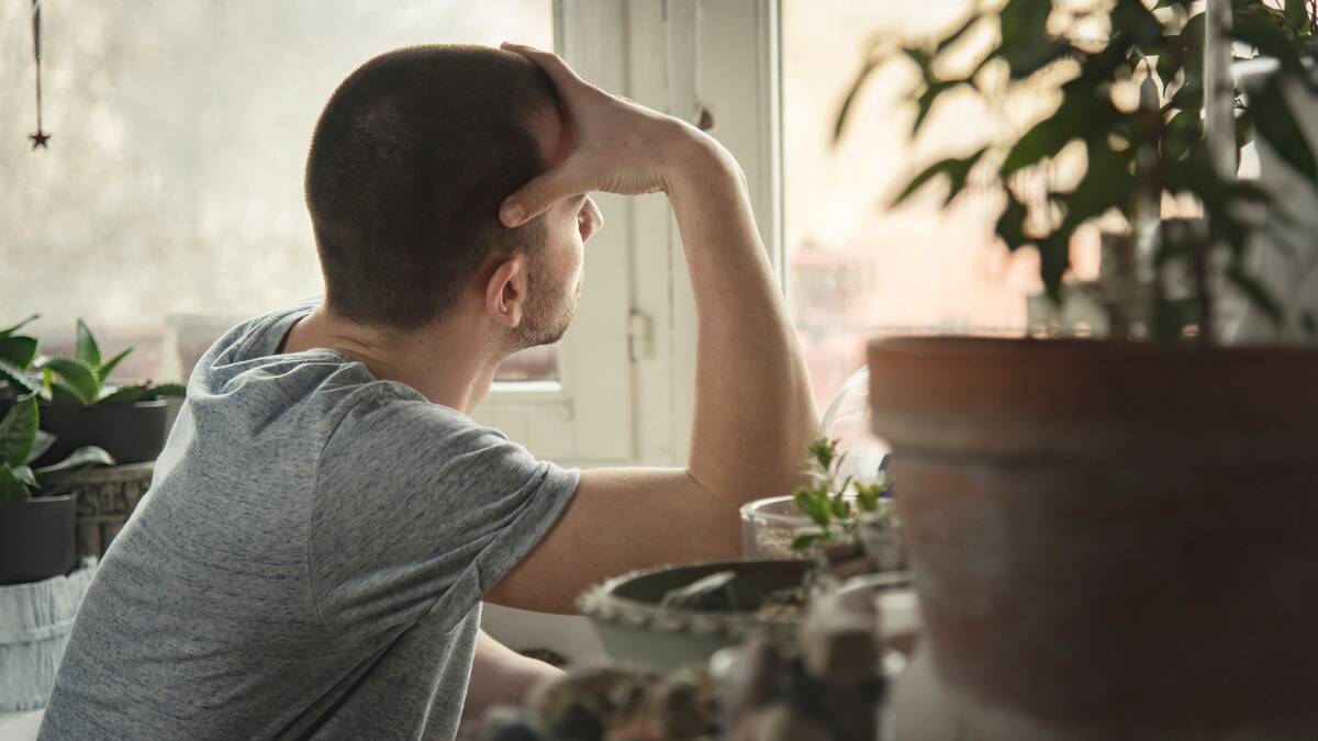 A man sitting by a window, one hand on his forehead, looking outside.