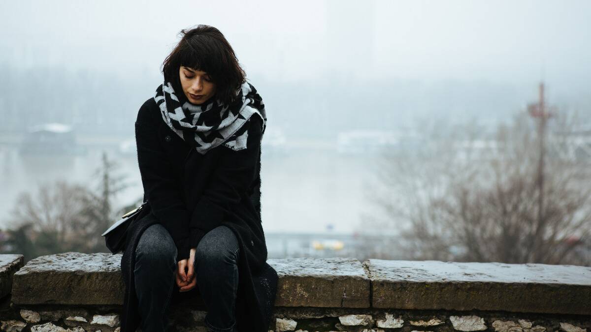 A woman sitting on a railing outside, looking down sadly.