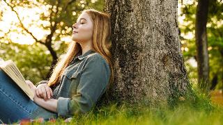 A woman sitting outside against a tree, book open on her lap, eyes closed as she smiles softly.