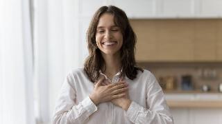 A woman smiling brightly with her eyes closed, both hands over her heart.