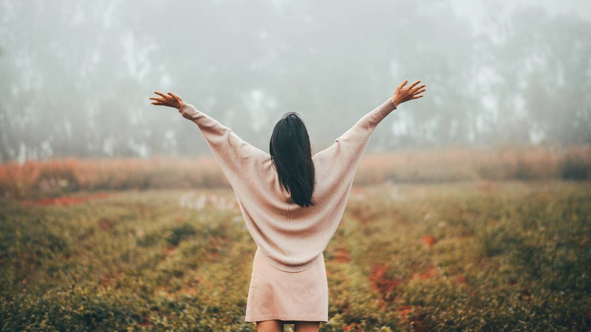 A woman standing outside among fog and flowers, arms up and outstretched, facing away from the camera.