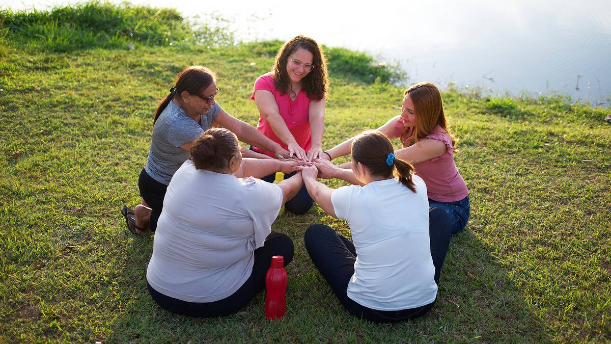 A group of women in a park sitting in a circle, all reaching their hands toward the center.