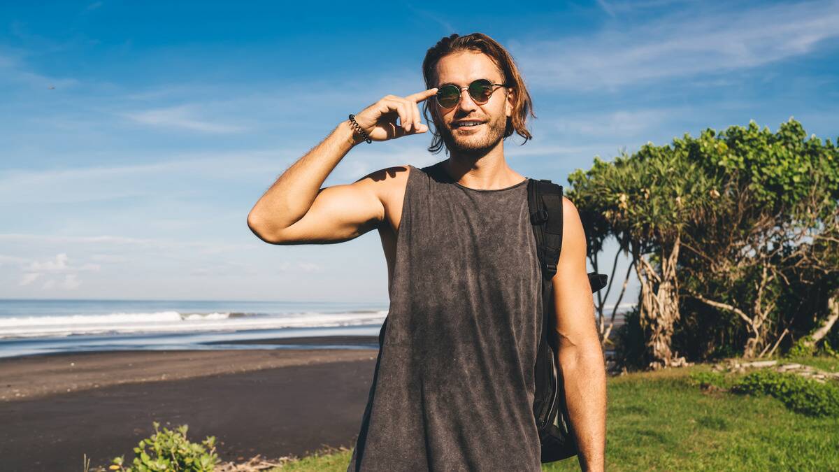 A man standing outside by the water, smiling and putting his fingers to the arm of his sunglasses.