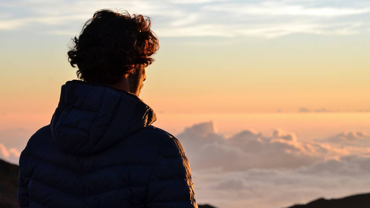 A man atop a mountain, above the cloud line, facing the just-rising sun.