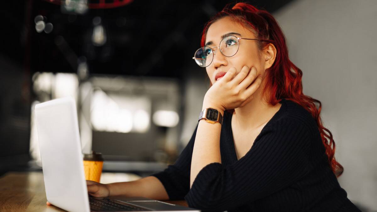 A woman sitting at her laptop in a cafe, resting her chin in her hand, looking upward with a thoughtful expression.