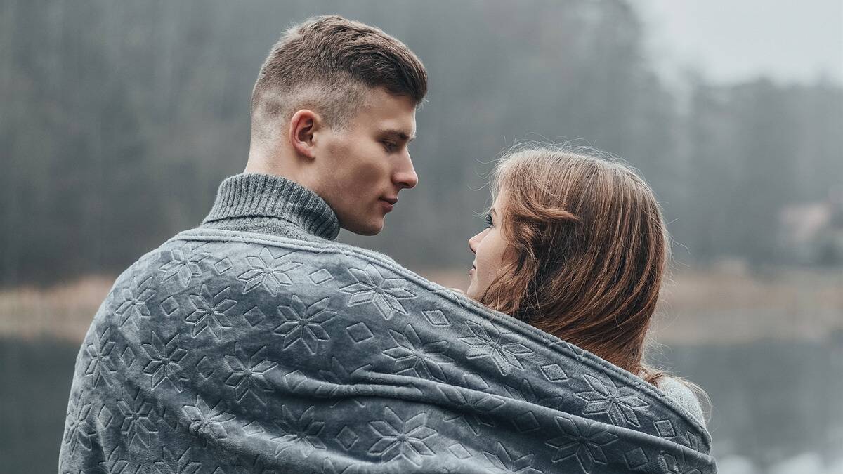 A couple standing outside, a blanket around their shoulders, looking at one another.