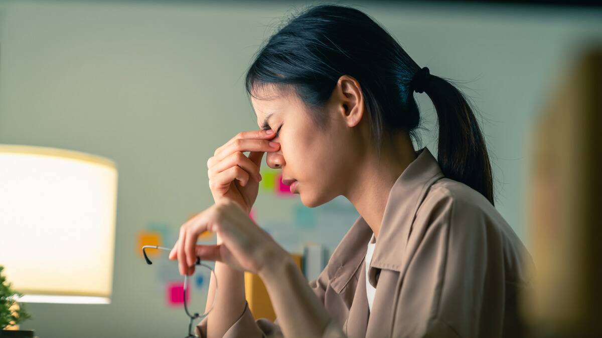 A profile shot of a woman holding her glasses and pinching the bridge of her nose, looking frustrated.