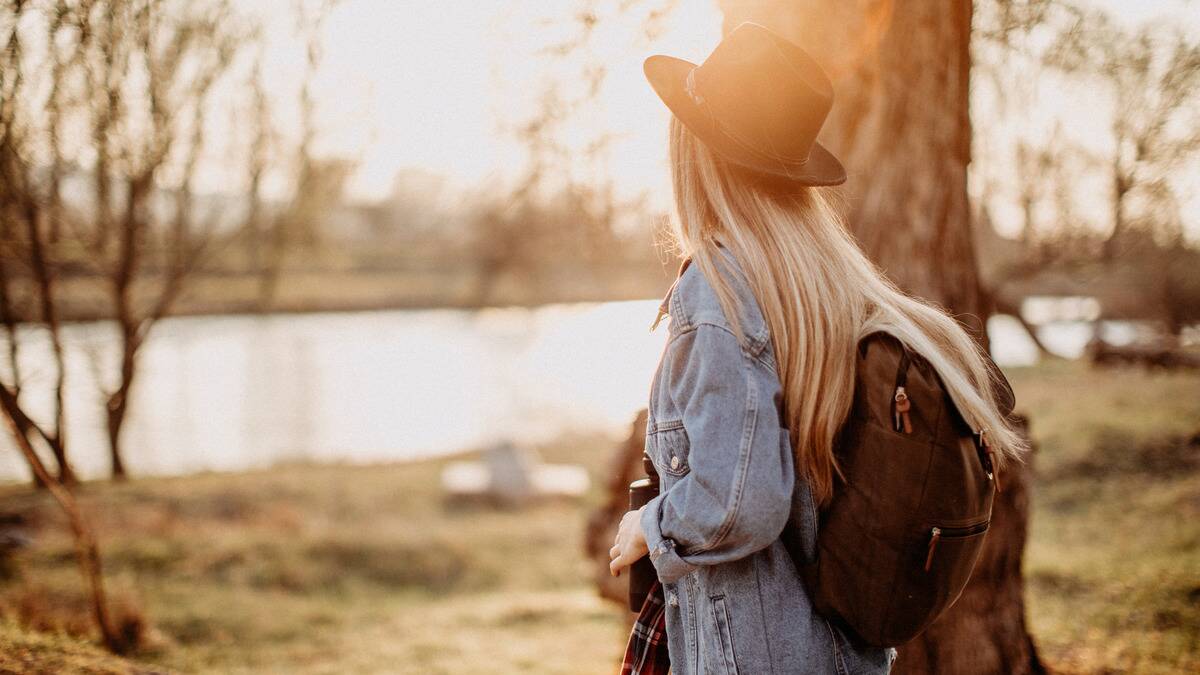 A woman with a backpack walking through a park, facing away from the camera, the sun shining behind her.