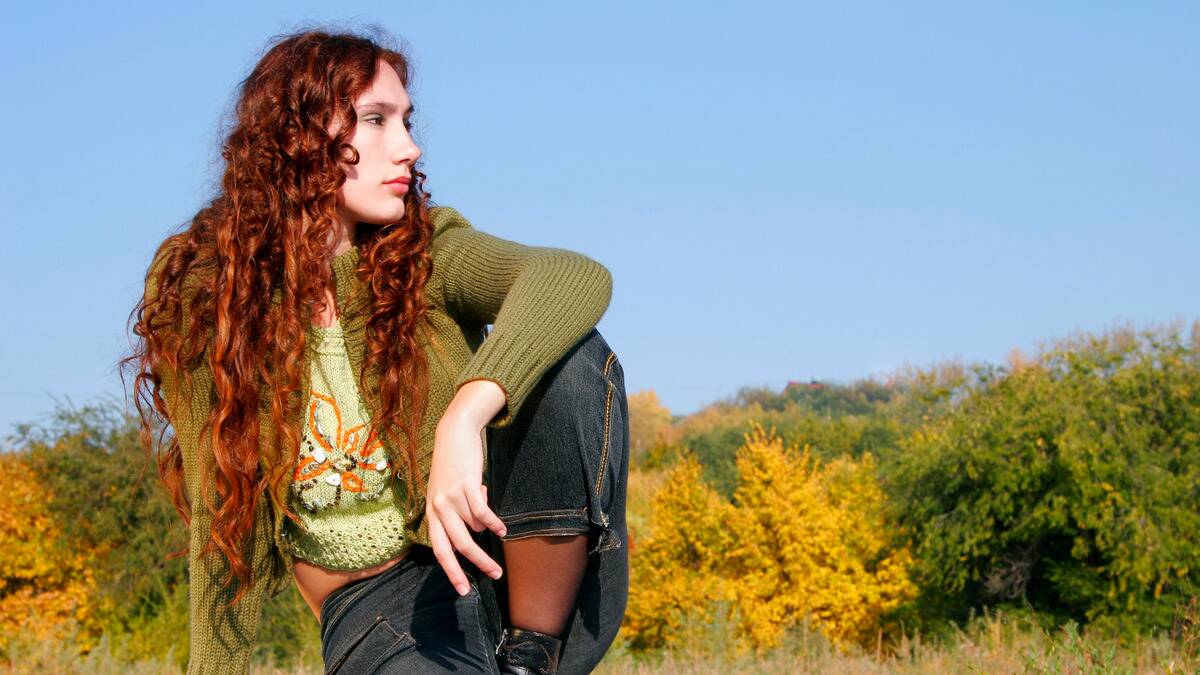 A woman sitting outside, one knee hiked up, her elbow resting on it as she looks off to the side.