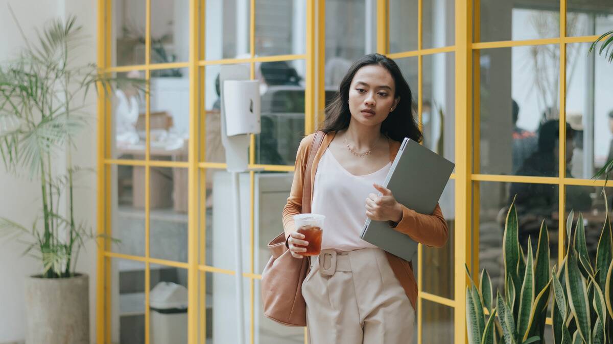 A business woman walking out of a meeting room with a laptop in one hand and an iced coffee in the other, looking tired.