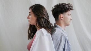 A man and woman standing back to back in front of a white backdrop, both looking melancholic.