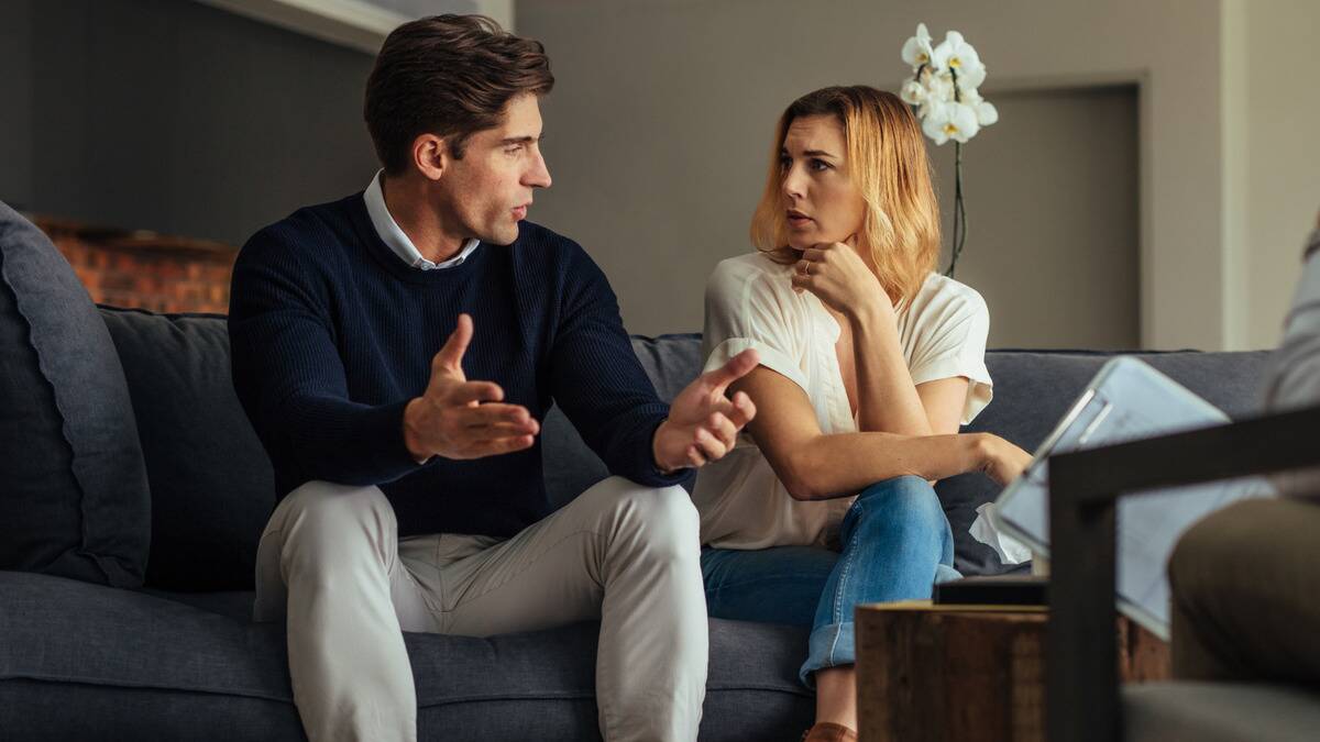 A couple looking incredulously at each other as they sit on a couch.