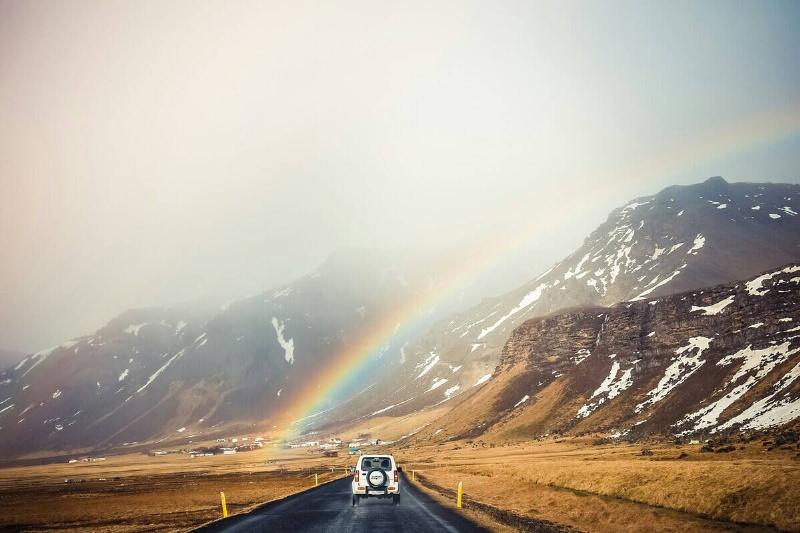 A faint rainbow touching the ground right next to a road in front of some mountains, a car driving down it.