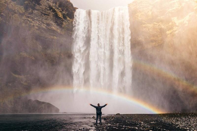 A man standing below a waterfall that has some rainbows stretching across it.