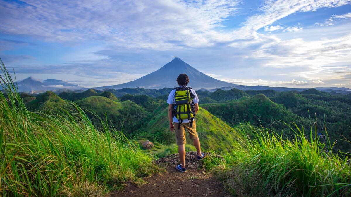 A man standing before a hilly range with a mountain in the background, facing away from the camera, one leg propped up.