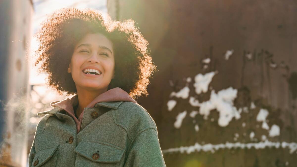 A woman smiling with her eyes closed as she stands outside in the sun.