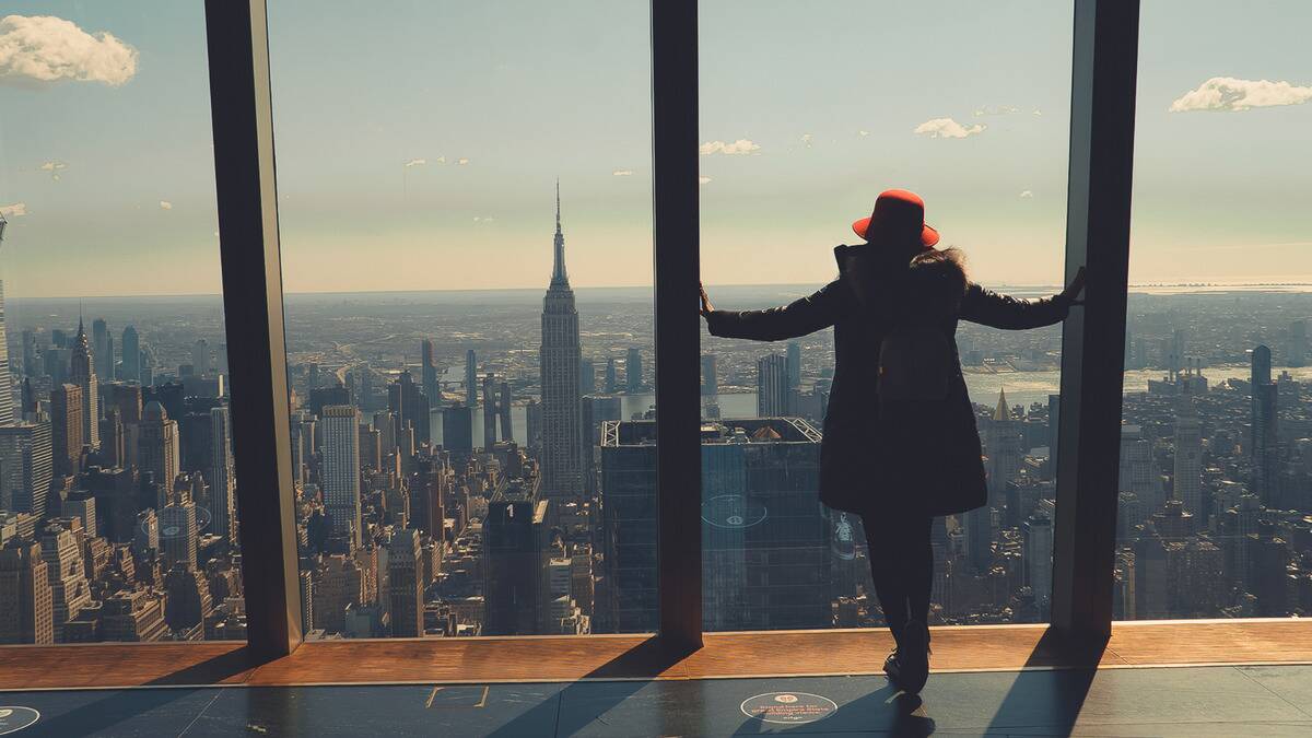 A woman standing in front of the large windows of a skyscraper floor, looking out at the cityscape before her.