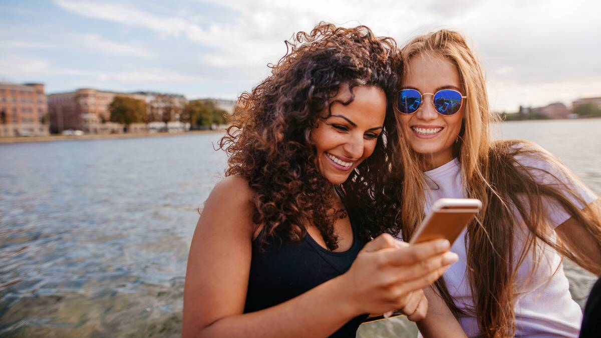 Two women standing outside by the water, both smiling as they lean into one another and look at one of their phones.