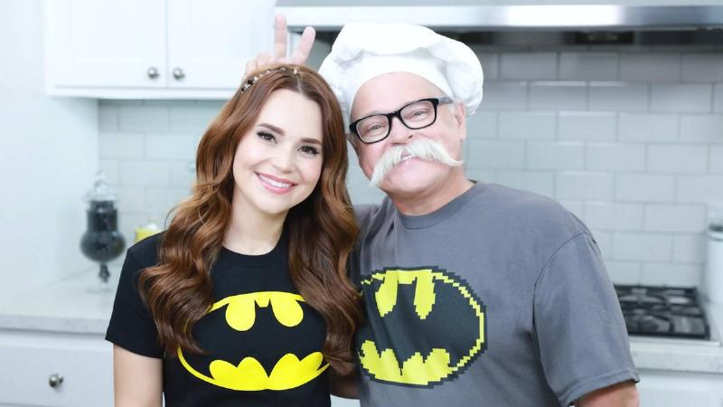 Rosanna standing next to her dad, who's wearing a chef's had and making bunny ears behind Rosanna's head with his fingers.