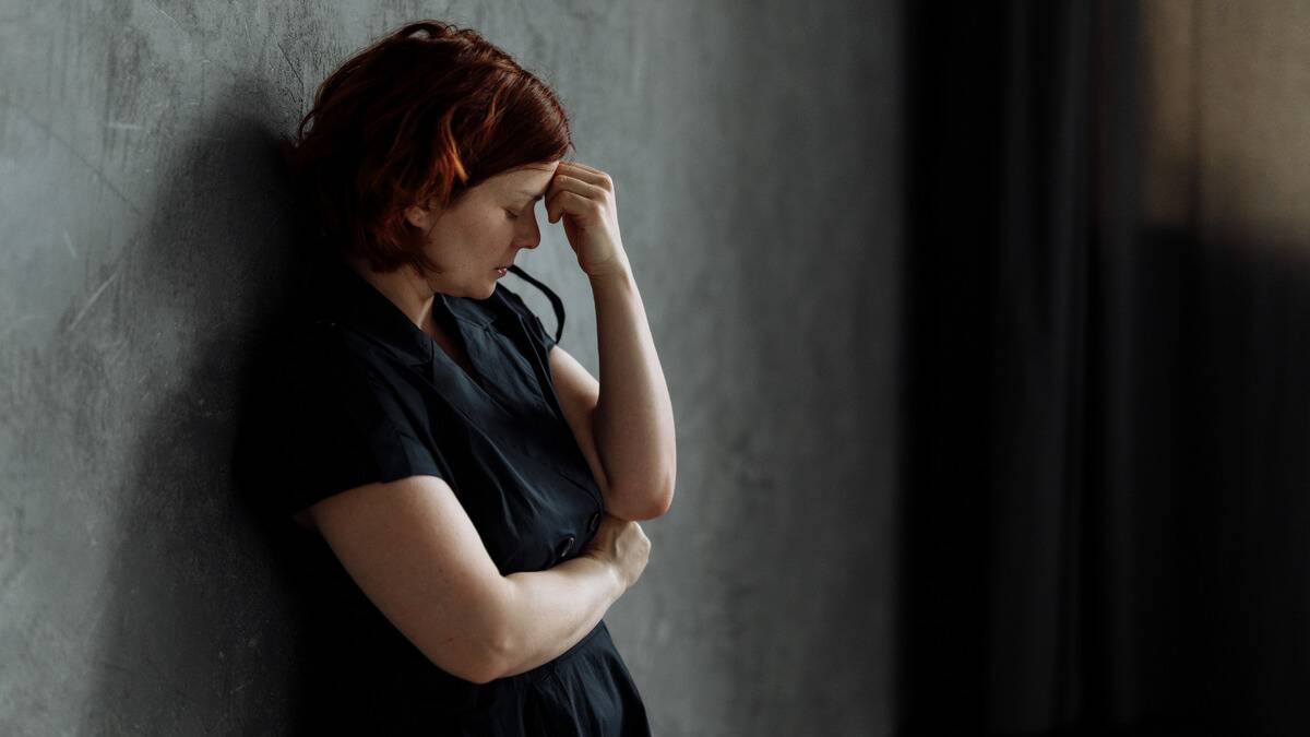 A woman standing with her back against a wall, hand to her forehead, looking frustrated.