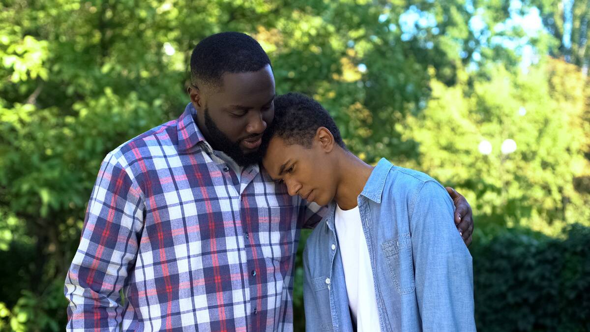 A teenage son leaning his head on his dad's shoulder, looking sad or guilty. His dad is comforting him with a hand on his shoulder.