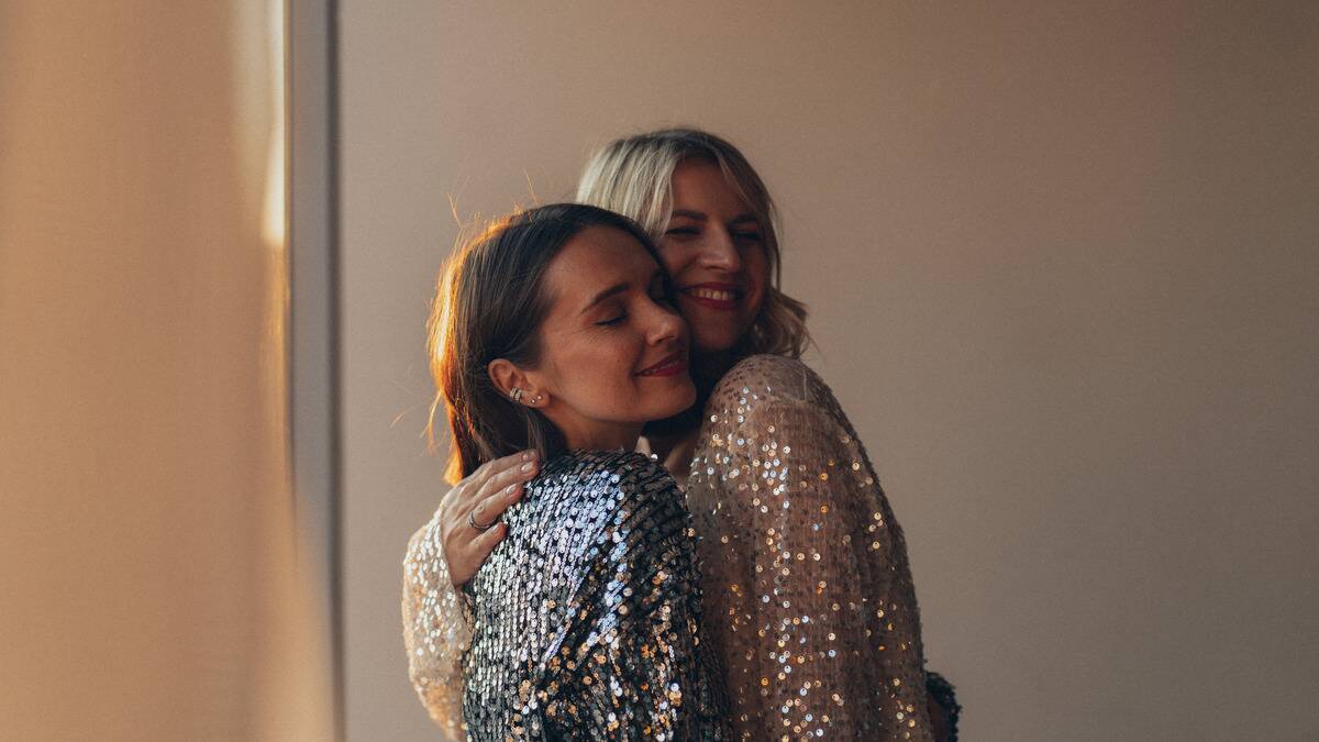 Two friends in sparkly, sequined dresses hugging, both smiling with their eyes closed.