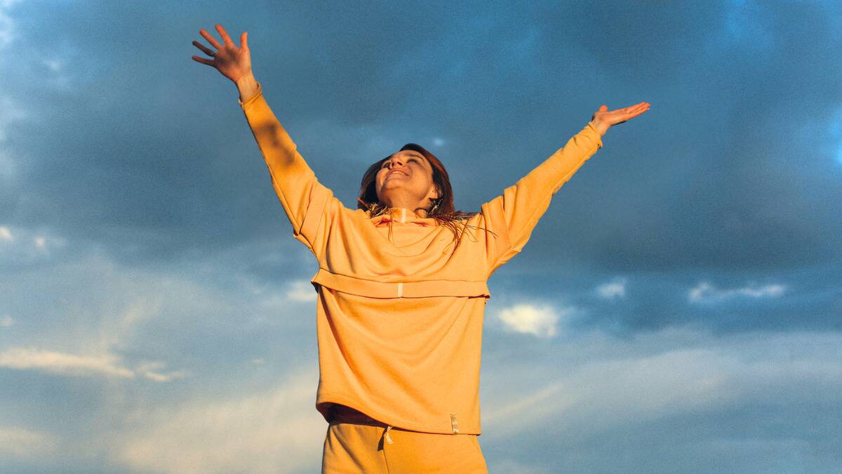 A woman standing beneath a cloudy blue sky, arms in the air, looking upward, smiling.