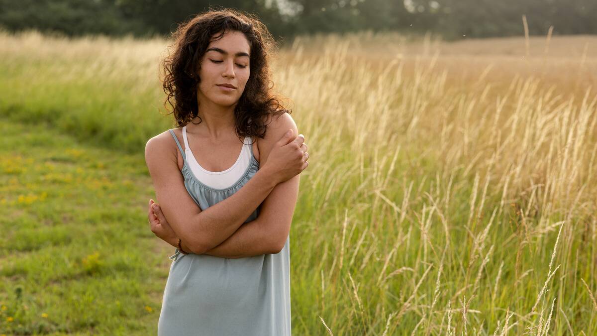 A woman standing in a field of tall grass with her eyes closed and her arms around herself, looking peaceful.