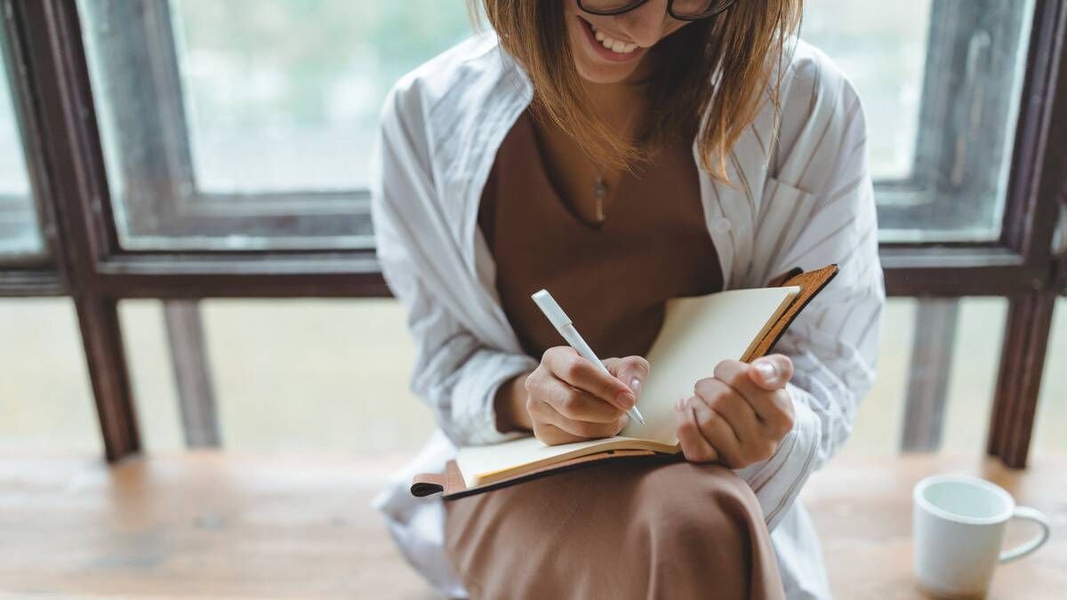 A woman smiling as she writes in an open journal that's propped up in her lap.