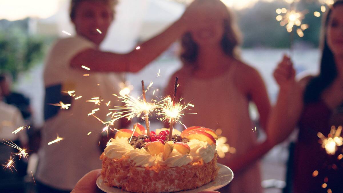 A cake with sparklers on top in the foreground. In the background, a woman is smiling as a man covers her eyes with his hand.