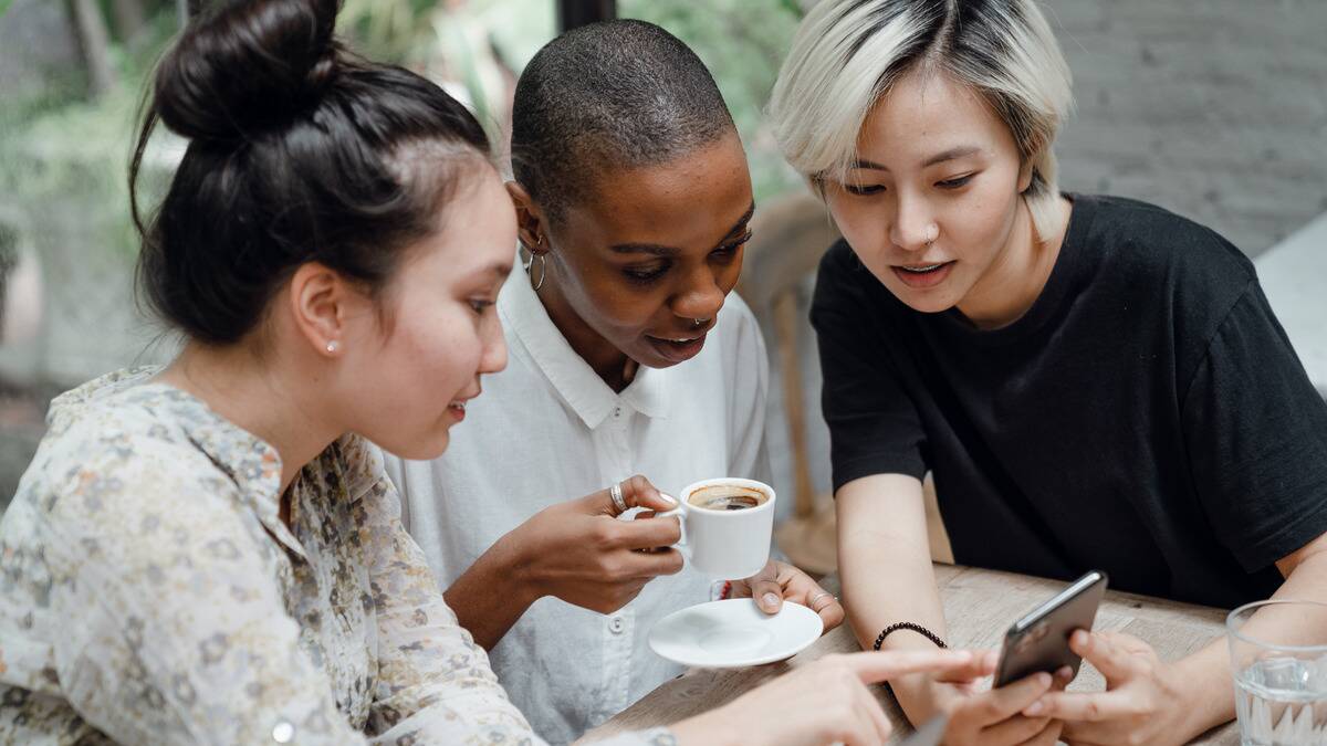 Three friends sat at a cafe table, two of them showing the third something on one of their phones.