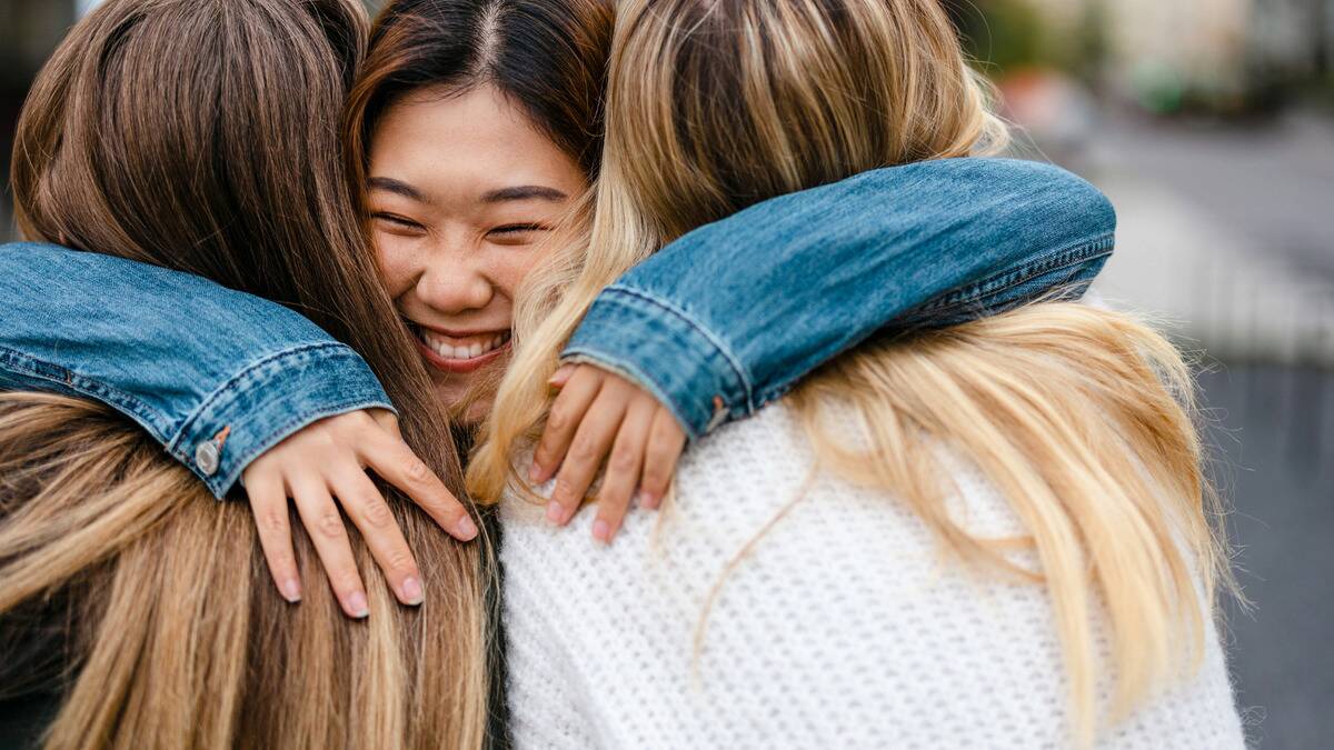 One girl being hugged by two of her friends, her smiling face seen between her friends' shoulders.